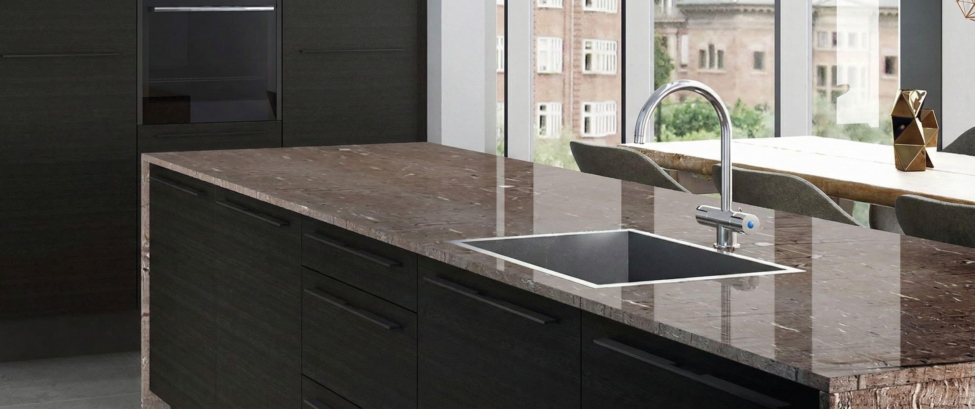 Kücheninsel mit schwarzer Front und glänzender Granit-Arbeitsplatte aus Naturstein; eckige Unterbauspüle mit hoher Chrom-Armatur vor großer Fensterfront und Essplatz im Hintergrund