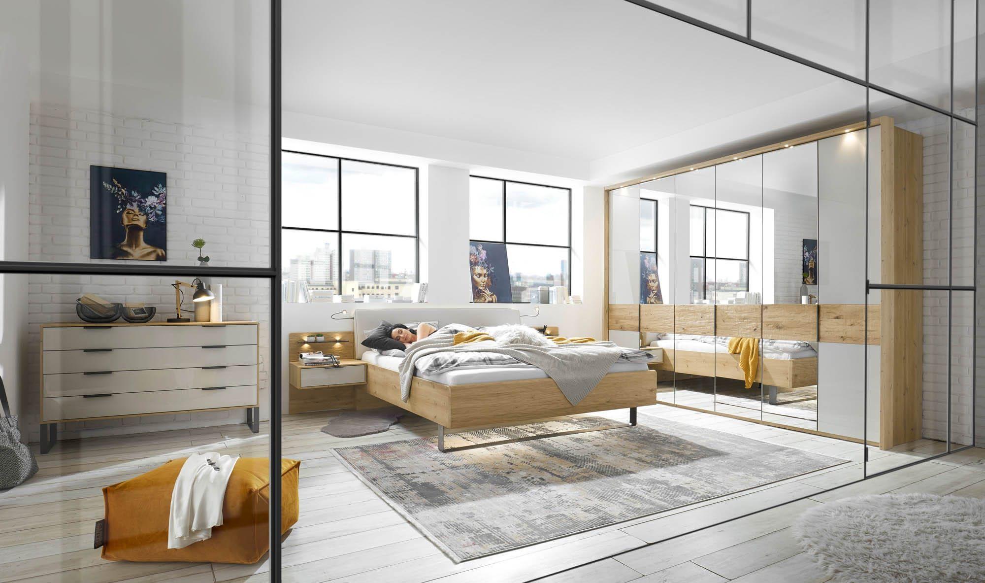 Calpe Calina Schlafzimmer-Programm in Eiche und Weiß mit Bett, Nachttischen mit integrierter Beleuchtung, Spiegelschrank und Kommode in modernem Loft mit Industriefenstern und Stadtblick