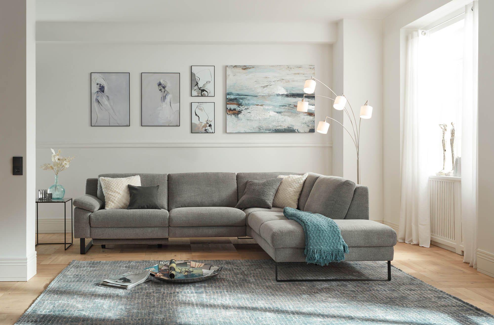 Modernes, helles Wohnzimmer mit großer grauer Eckcouch auf blau-grauem Teppich, passenden Kissen und Decke, Bildergalerie an der Wand und Stehlampe am Fenster