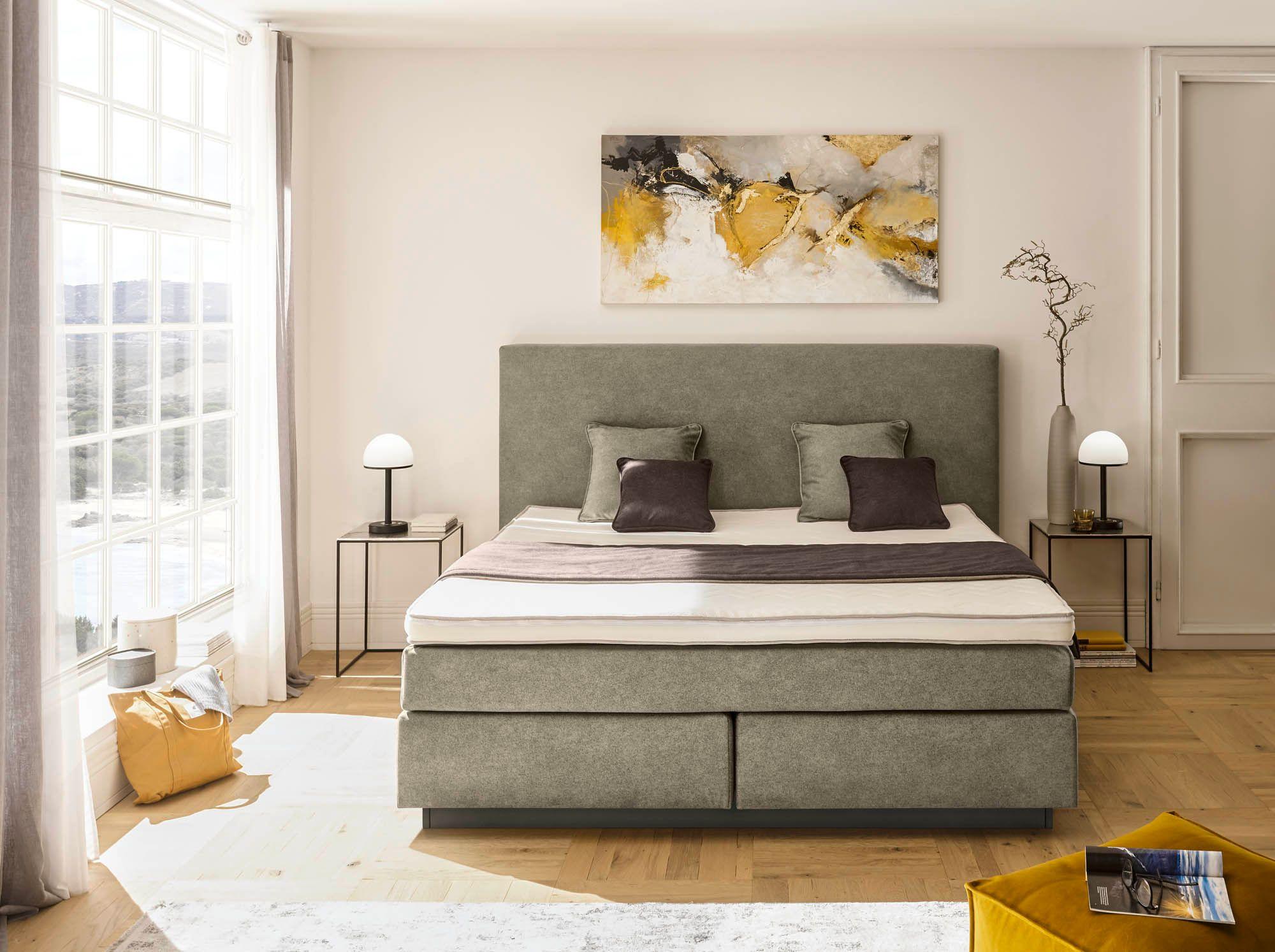 Frankenstolz Boxspringbett grau mit schlichtem Kopfteil und taupefarbenen Kissen in lichtdurchflutetem Schlafzimmer mit modernem Kunstwerk
