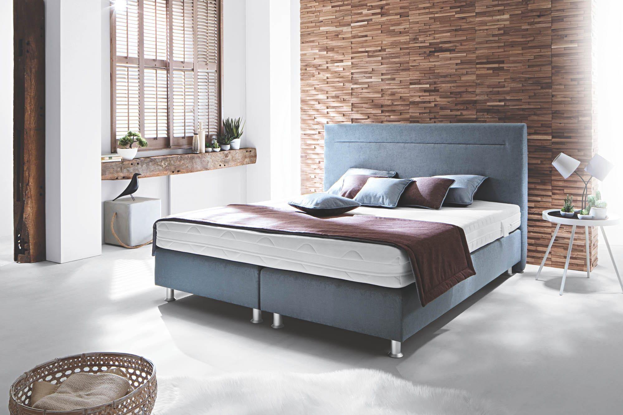 Frankenstolz Classic Express Boxspringbett in Graublau mit weißer Matratze und brauner Tagesdecke, dekoriert mit farblich abgestimmten Kissen in einem modernen Schlafzimmer mit Holzpaneel-Wand