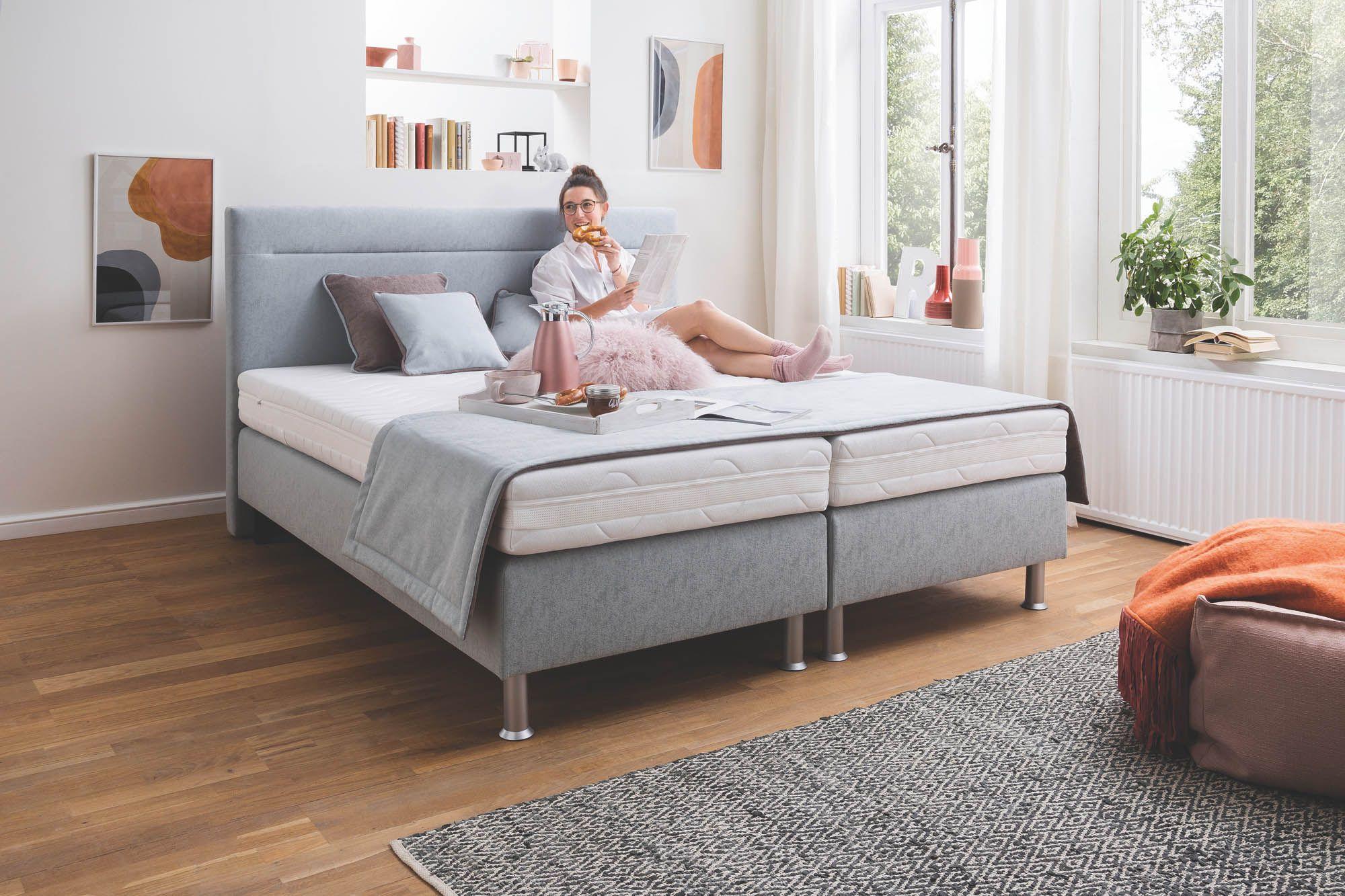 Frankenstolz Classic KT101 Boxspringbett in Hellgrau mit gepolstertem Kopfteil – Frau genießt entspannt Frühstück im Bett in hellem, modernem Schlafzimmer mit Holzboden