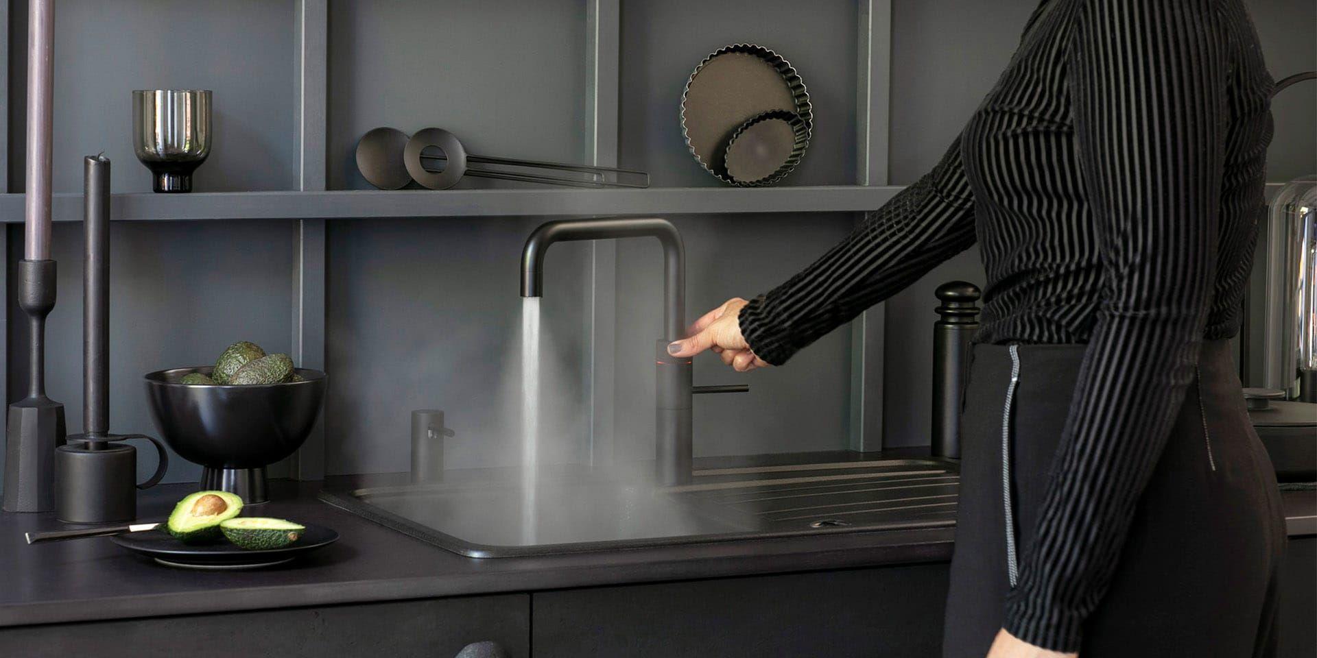 Quooker Fusion Square Black – schwarzer Kochendwasserhahn in moderner Küche; Person zapft heißes Wasser am Spülbecken, sichtbarer Dampf, dunkle Arbeitsfläche mit Avocado und Küchenaccessoires