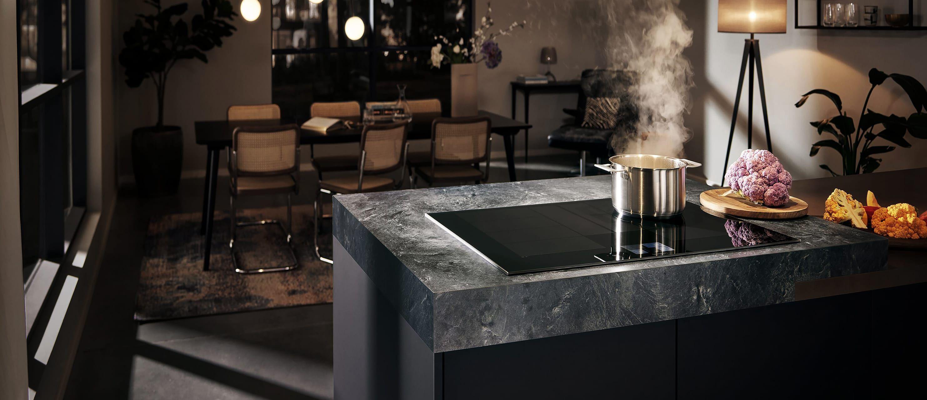 Dunkle Kücheninsel mit Arbeitsplatte in Steinoptik und flächenbündigem Siemens Induktionskochfeld; dampfender Kochtopf und Blumenkohl auf Schneidebrett in modernem Essbereich bei warmem Licht