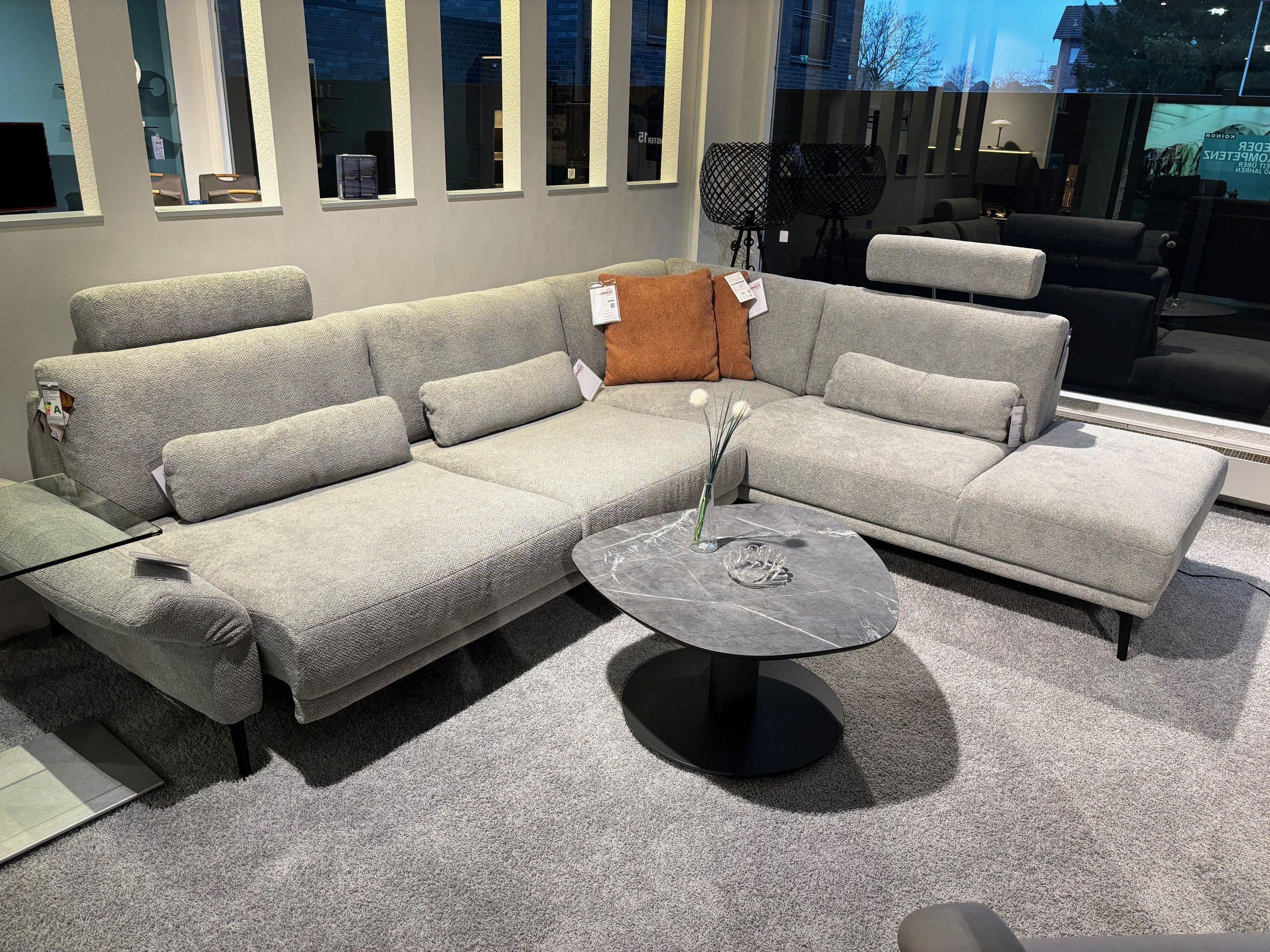 Hellgraue Polster-Eckgarnitur mit offener Récamiere und flexiblen Armlehnkissen, dazu runder Marmor-Couchtisch im Showroom.